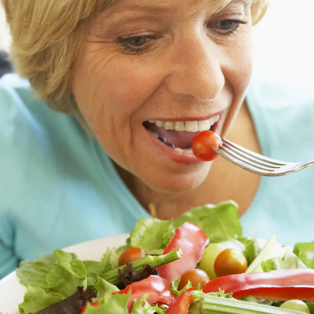 Mujer de mediana edad que come una ensalada con mucho afán y sonriendo, está en primer plano