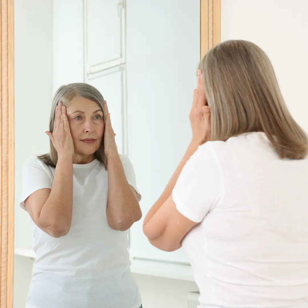 Mujer de mediana edad que se mira al espejo con las manos a ambos lados de la cara como si no se gustase.